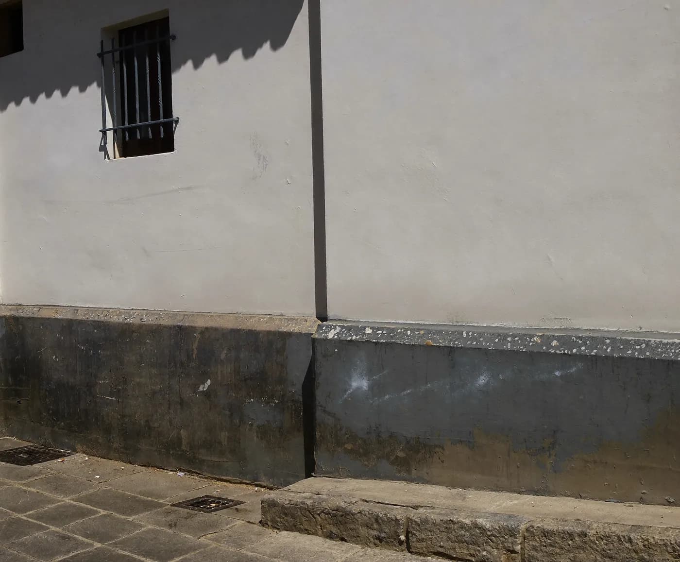 Fachada encalada de una casa de una planta con ventana enrejada y un zócalo gris oscuro con manchas de humedad sobre la acera