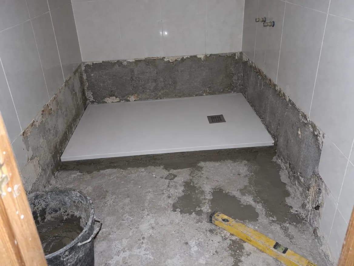 Hueco de una bañera retirada con un plato de ducha blanco extraplano colocado sobre mortero, la franja inferior de pared sin azulejo, un cubo y un nivel